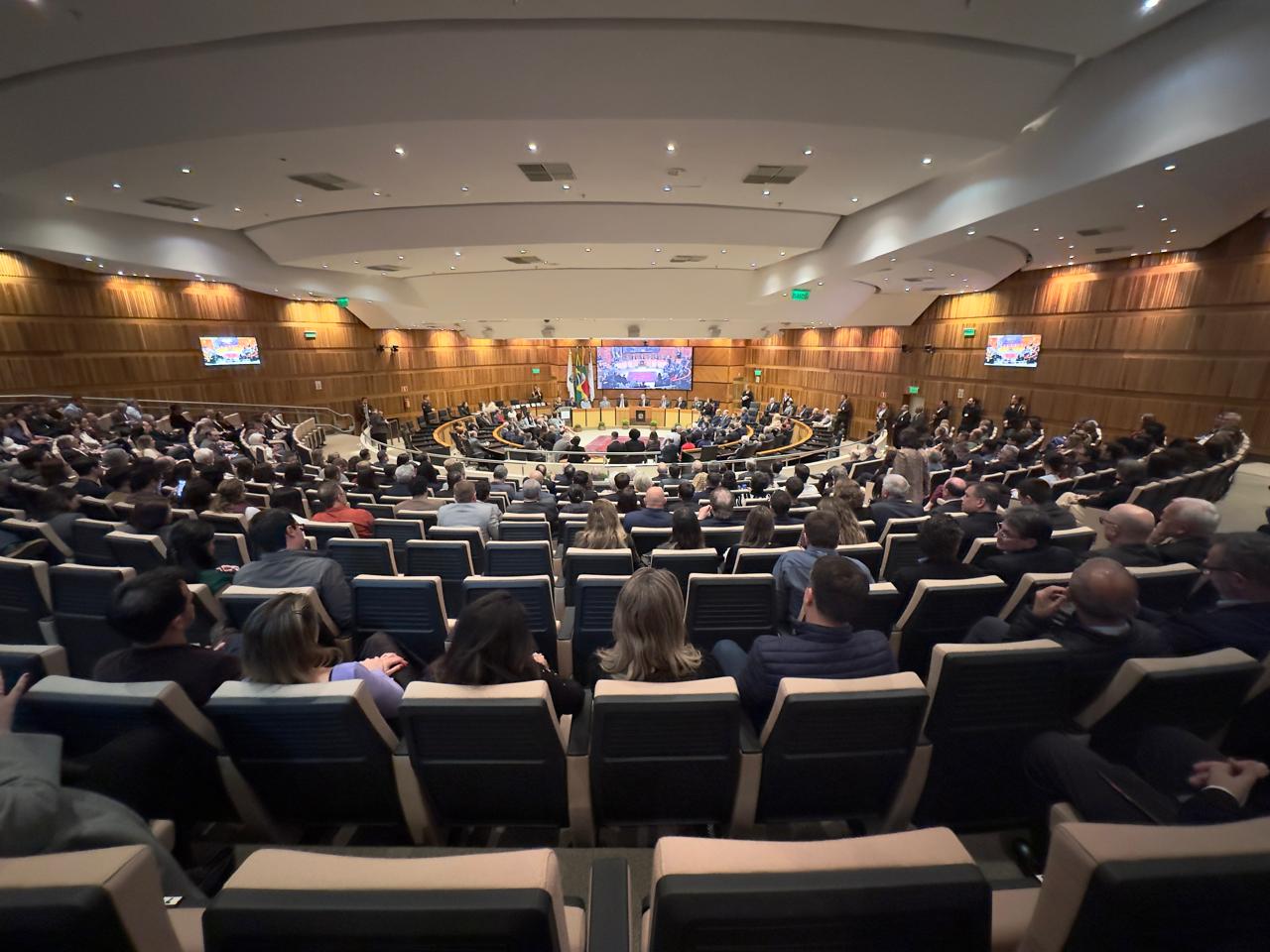 Foto do Plenário com público em primeiro plano e nave central com o telão ligado ao fundo.