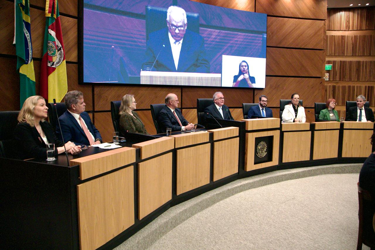Mesa de abertura da Semana da Conciliação Trabalhista, com autoridades e representantes sentados em um plenário formal. Ao fundo, bandeiras do Brasil e do Rio Grande do Sul. Um telão exibe o discurso de abertura de um dos integrantes, com intérprete de Libras ao lado. O ambiente tem decoração em madeira e brasão oficial à frente da mesa.