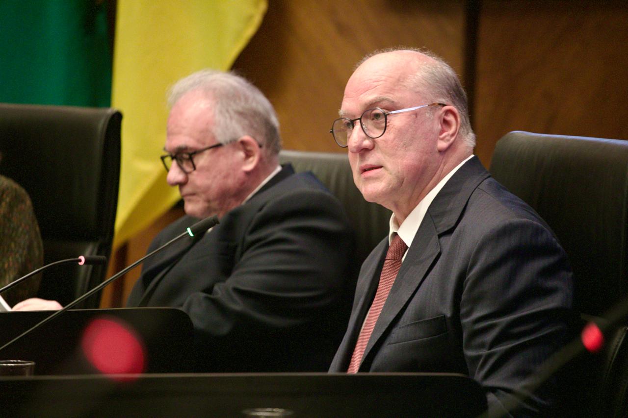 Presidente do Tribunal Regional do Trabalho da 4ª Região (TRT-RS), desembargador Ricardo Martins Costa, durante a mesa de abertura da Semana da Conciliação Trabalhista. Ele está sentado à frente de um microfone, usando terno escuro, camisa branca e gravata vermelha. Ao fundo, aparece parcialmente o vice-presidente do TST, ministro Maurício Godinho Delgado, com bandeiras ao fundo e ambiente de plenário solene.
