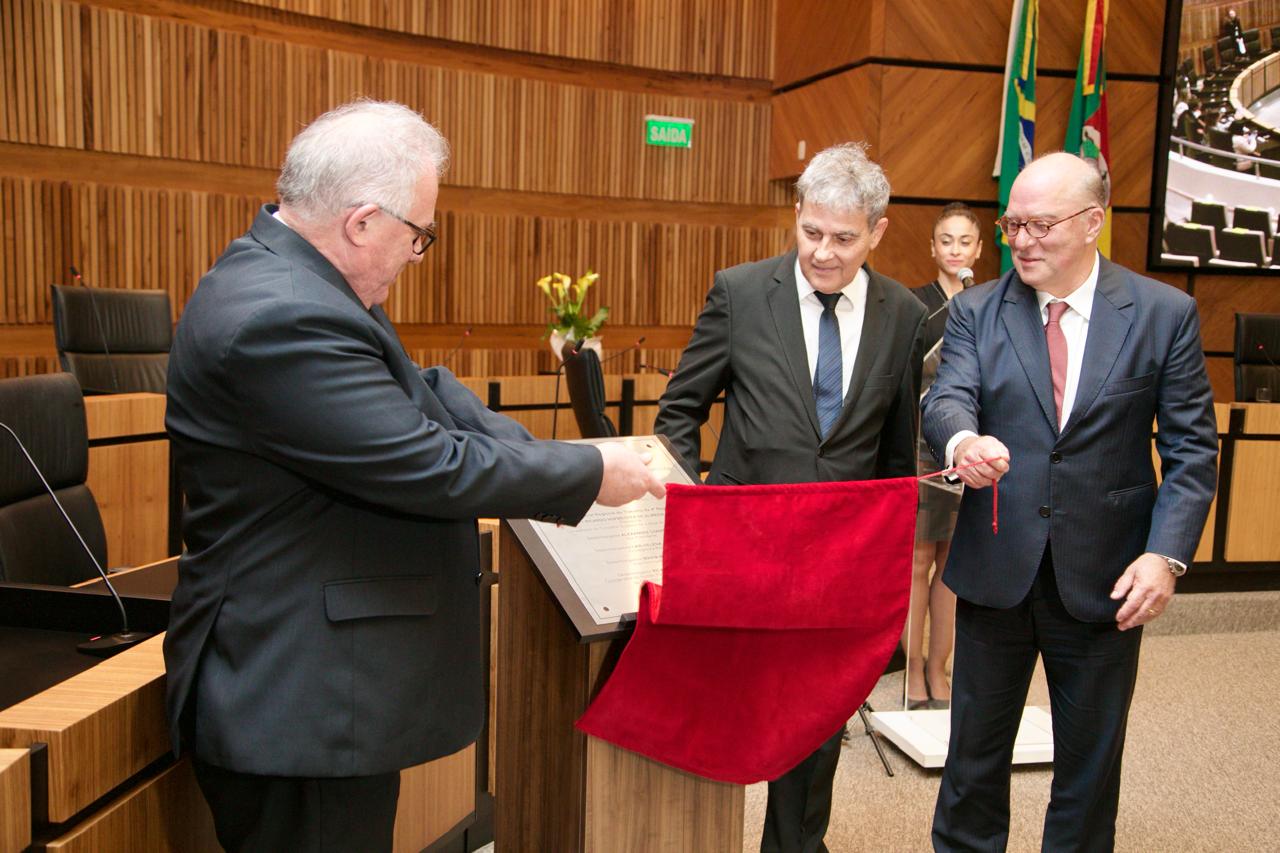 Os desembargadores Godinho, Ricardo Carvalho Fraga e Ricardo Martins Costa realizam o descerramento de uma placa comemorativa durante a cerimônia de abertura da Semana da Conciliação Trabalhista no plenário do TRT-RS. O ato simbólico ocorre diante de um púlpito de madeira, com uma cortina vermelha sendo puxada. Ao fundo, aparecem bandeiras e parte da estrutura moderna do plenário, com painéis de madeira.
