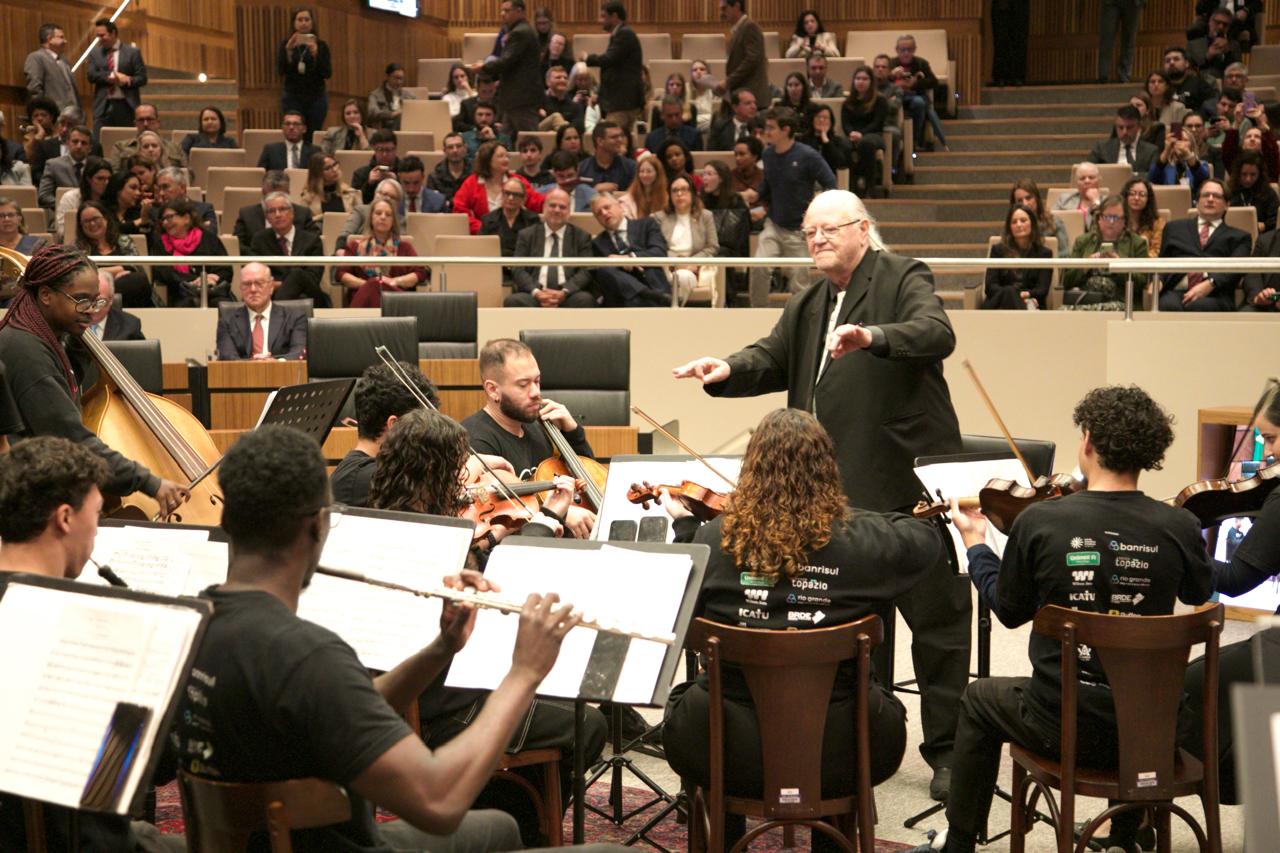  Maestro rege orquestra formada por jovens músicos durante apresentação no plenário Milton Varela Dutra. Ao fundo, o auditório está lotado, com o público sentado nas arquibancadas assistindo atentamente.