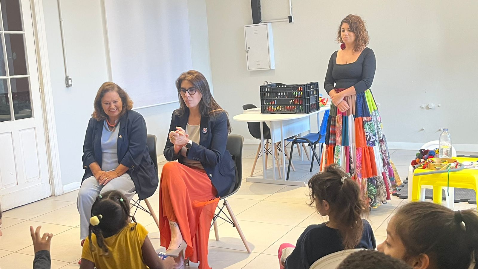 Três mulheres adultas interagem com um grupo de crianças em uma sala de aula. Duas delas estão sentadas em cadeiras, sorrindo e conversando com as crianças, enquanto a terceira está em pé, observando. A mulher sentada à esquerda usa um conjunto azul-claro com blazer azul-marinho, a do centro veste blazer escuro e uma saia longa laranja. A mulher em pé usa uma blusa preta e uma saia longa colorida em retalhos. À frente, várias crianças pequenas assistem atentamente. Ao fundo, há mesas com brinque