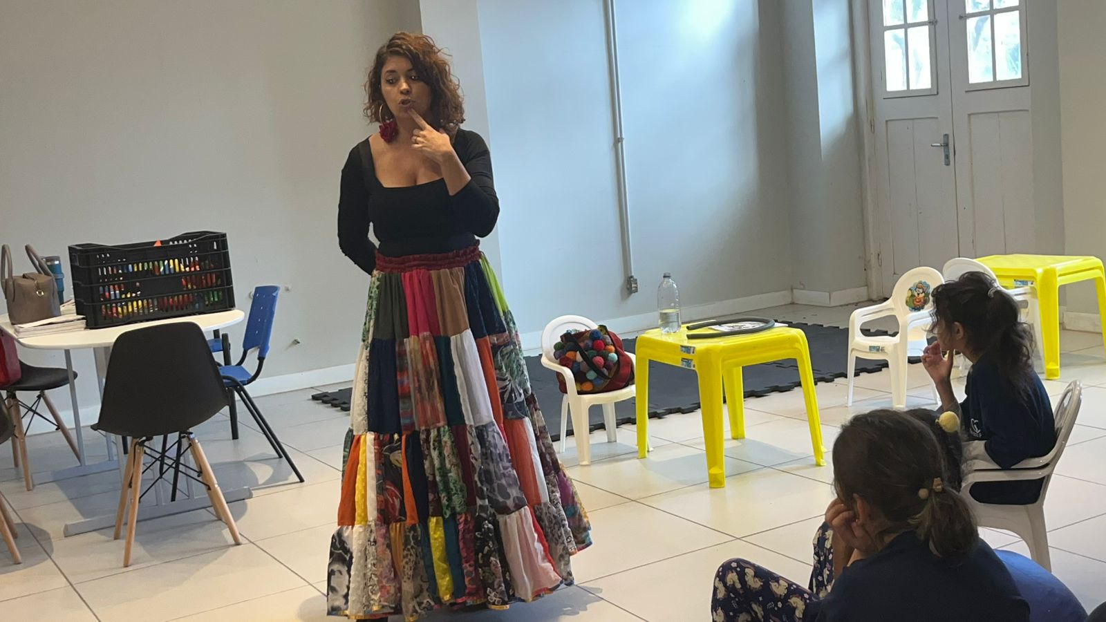 Mulher com blusa preta de mangas longas e saia longa colorida feita de retalhos fala para um pequeno grupo de crianças em uma sala de atividades. Ela está em pé, com uma das mãos no rosto, sugerindo que está contando ou explicando algo. À frente, duas meninas sentadas a observam atentamente. No fundo, há mesas e cadeiras infantis amarelas e brancas, uma garrafa de água e materiais didáticos, como brinquedos e livros. O ambiente é claro, com paredes brancas e janelas que deixam entrar luz natural