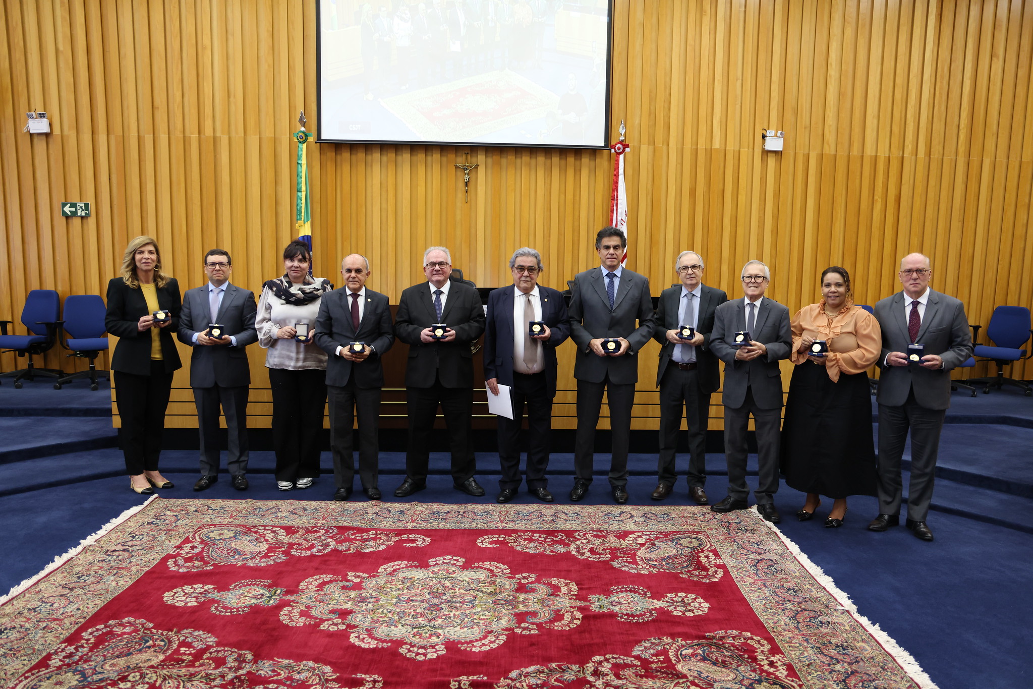 Dois homens em trajes formais, um de terno azul escuro e outro de terno cinza, posando em uma sala com paredes de madeira amarela. O homem à esquerda entrega um certificado ao homem à direita, que segura uma medalha. O certificado menciona "Ricardo Martins-Costa" e a data "23 de maio de 2025".