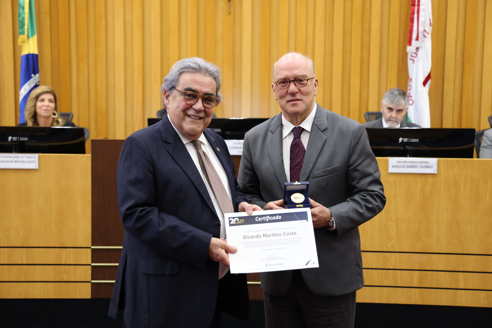 Dois homens em trajes formais, um de terno azul escuro e outro de terno cinza, posando em uma sala com paredes de madeira amarela. O homem à esquerda entrega um certificado ao homem à direita, que segura uma medalha. O certificado menciona "Ricardo Martins-Costa" e a data "23 de maio de 2025".
