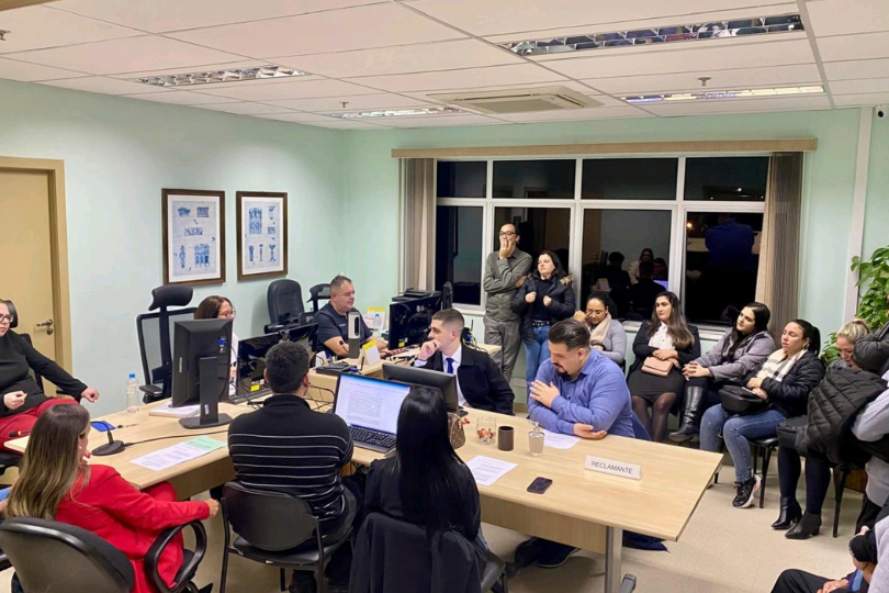 Juízas e estudantes na sala de audiência. Eles estão sentados em volta de ume mesa de cor bege claro com monitores de computadores sobre o móvel.