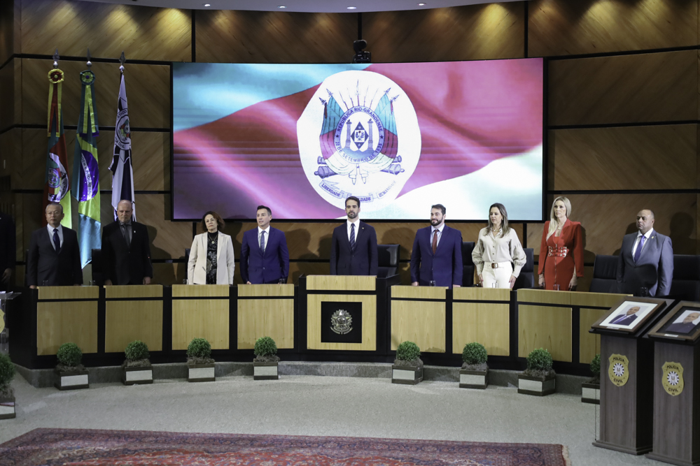 Nove pessoas aparecem de pé, na mesa oficial. Atrás deles, o telão mostra a bandeira do Rio Grande do Sul