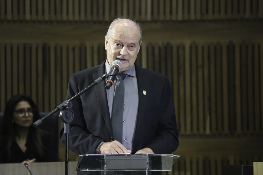 Heraldo é e grisalho. Usa terno preto e camisa cinza com gravata cinza. Ele está de pé no púlpito, falando e gesticulando.