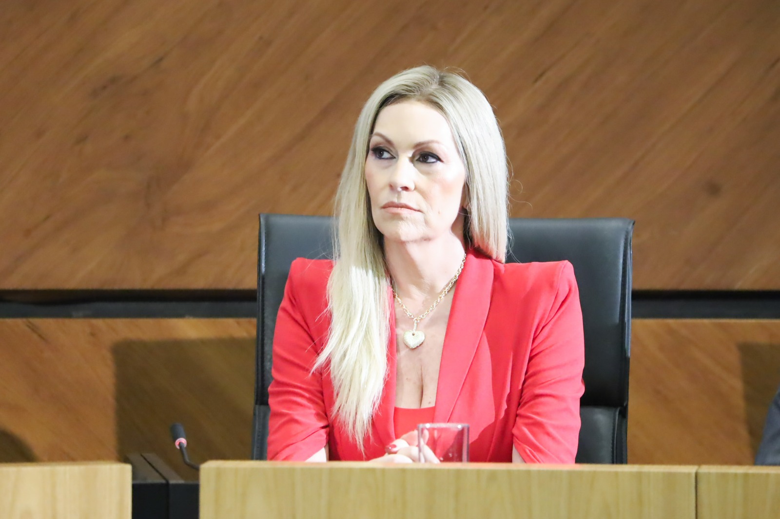Adriana é branca, tem cabelo loiro liso e comprido. Está olhando para esquerda da foto. Usa um tailler vermelho. Está sentada na mesa oficial.
