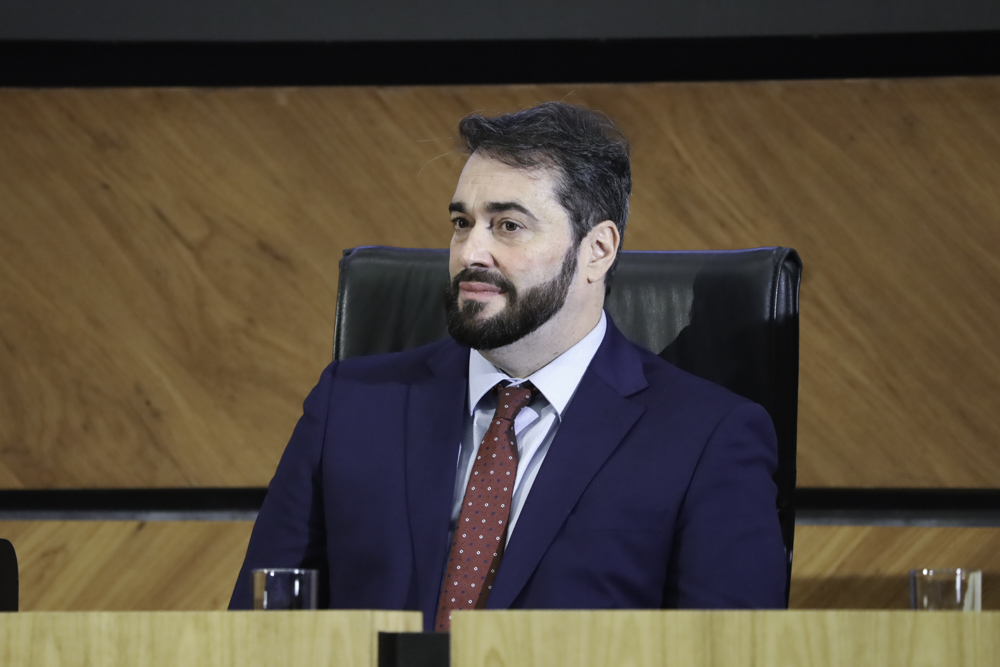 Alexandre é branco, tem cabelo curto escuro e barca escura. Usa terno azul marinho, camisa branca e gravata bordô. Está sentado na mesa oficial.