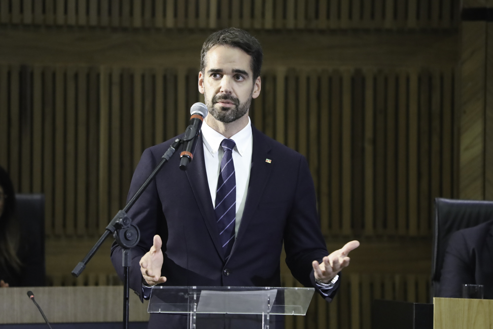 Eduardo é branco de cabelo castanho. Usa barba curta. Usa terno azul marinho, camisa branca e gravata azul. Está de pé no púlpito, fala e gesticular.