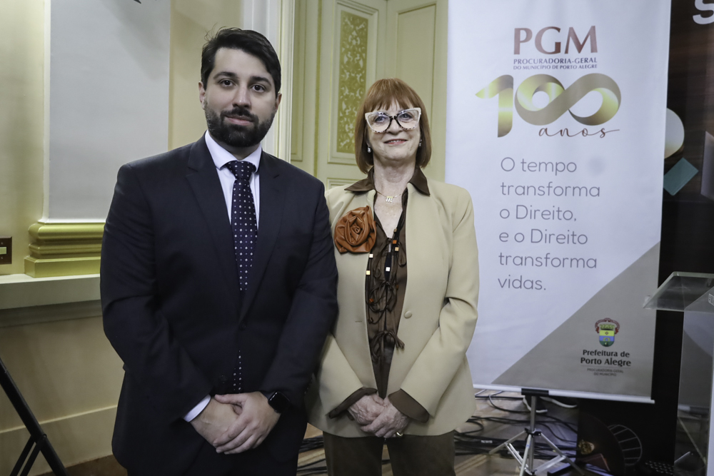 Jhonny é branco, de cabelo e barba escura. Veste terno azul marinho, camisa azul e gravata azul. Madalena é branca e ruiva. Usa blazer creme com decoração de flor laranja, e camisa e calça marrom. Usa óculos de aro grande. Estão de pé posando para a foto e sorrindo. No lado direito aparece um cartaz com o logo dos 100 anos da PGM e o texto "o tempo transforma o Direito. e o Direito transforma vidas.