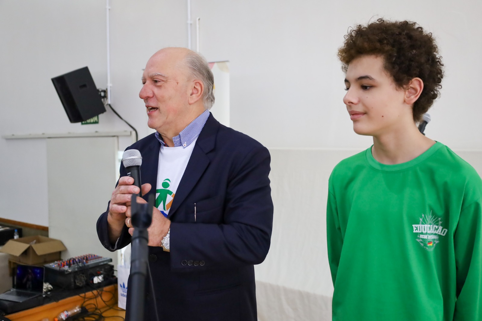 Des. Martins Costa falando ao microfone, ao lado de um jovem estudante.