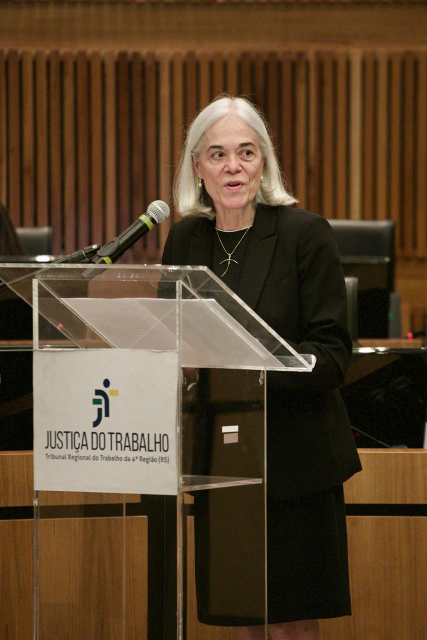 Maria Thereza é branca, de cabelos grisalhos longos e lisos Veste camisa, blazer e saia pretos. Ela está de pé e fala ao microfone sorrindo, olhando pra a direita. Em frente ao púlpito aparece o logo da Justiça do Trabalho.