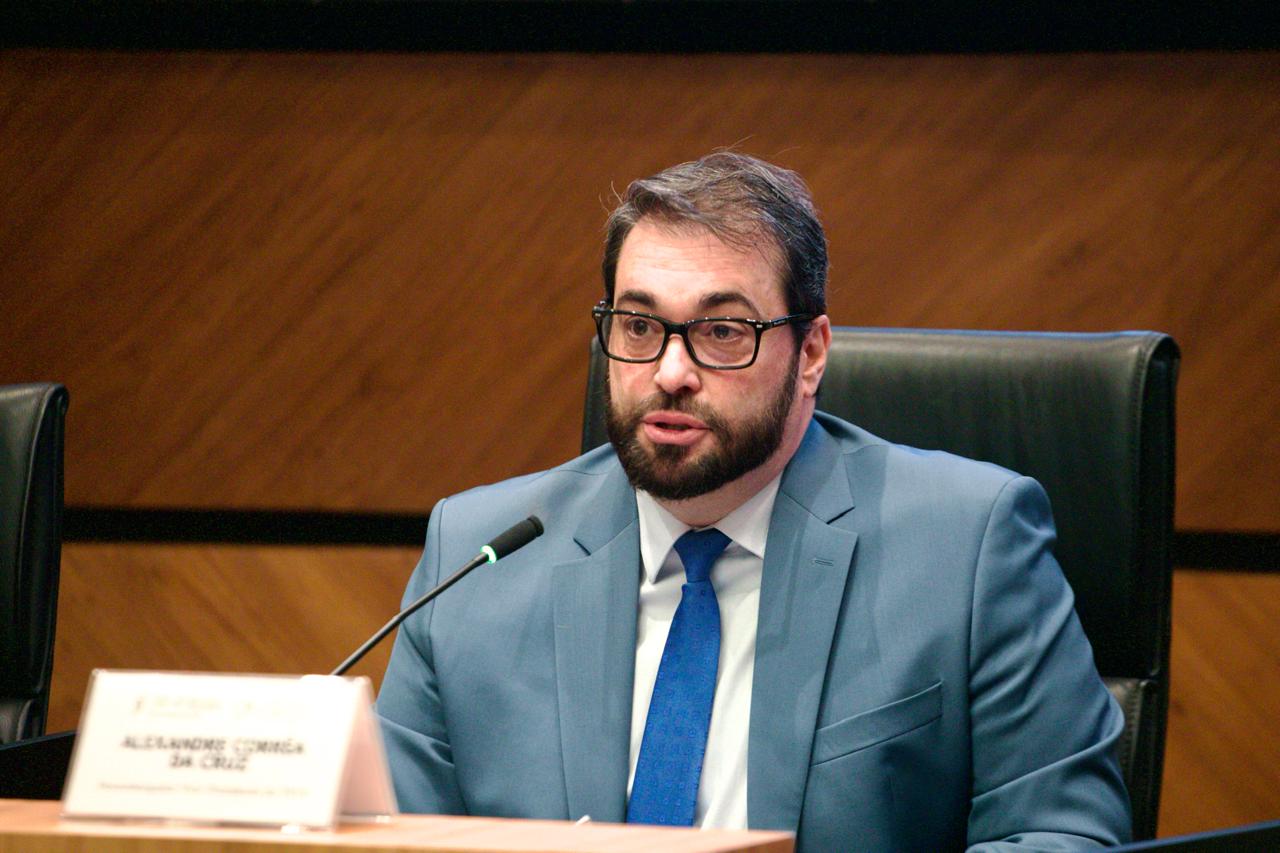 Alexandre é branco, usa barba e cabelo pretos. Veste blazer azul claro, camisa branca e gravata azul. Está falando ao microfone. Usa óculos de aro preto.