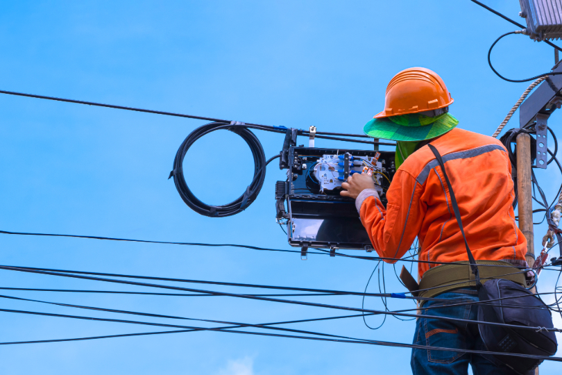  homem trabalha em instalações em poste elétrico. Ele está de costas e usa capacete e camisa cor de laranja e calça jeans