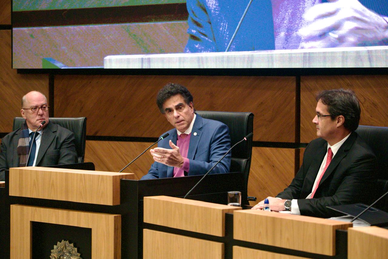 O presidente do TRT-RS, desembargador Ricardo Martins Costa, o corregedor-geral da Justiça do Trabalho, ministro Luiz Philippe Vieira de Mello Filho, e seu assessor Cláudio Gomes Carneiro participam de uma atividade no Plenário Milton Varela Dutra. Sentados à mesa, o ministro fala enquanto os outros escutam. Ao fundo, um telão exibe a transmissão do evento.
