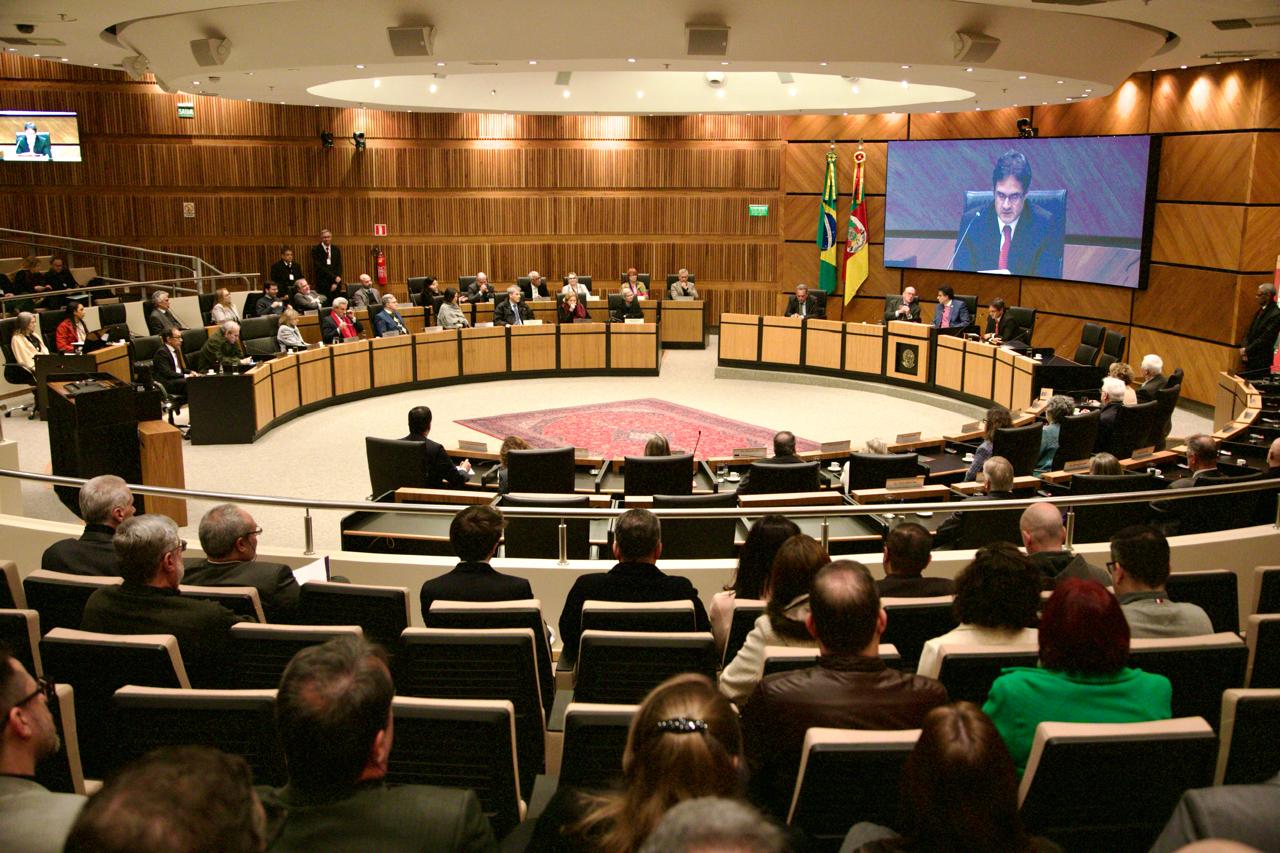 O Plenário Milton Varela Dutra, do TRT-RS, recebeu diversas autoridades e servidores em um evento que contou com transmissão ao vivo por telão. O espaço tem formato semicircular, com cadeiras distribuídas em diferentes níveis, proporcionando boa visibilidade para o público. Ao fundo, estão as bandeiras do Brasil e do Rio Grande do Sul, além de uma grande tela que exibe os discursos em tempo real.