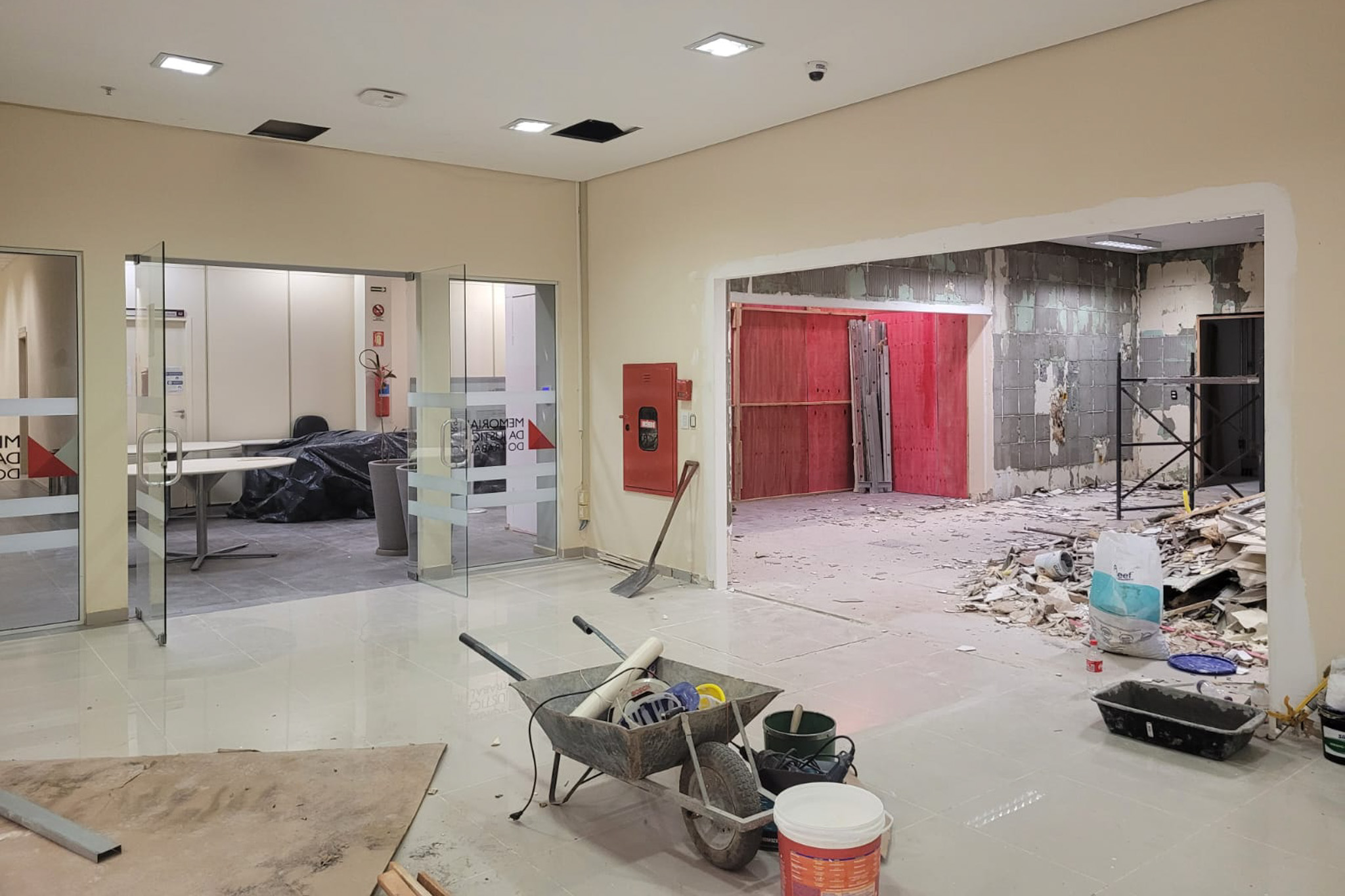 Sala em obras aparece um carrinho de mão em primeiro plano. ao fundo as portas de vidro estão abertas e à direta uma outra sala está em obras também.