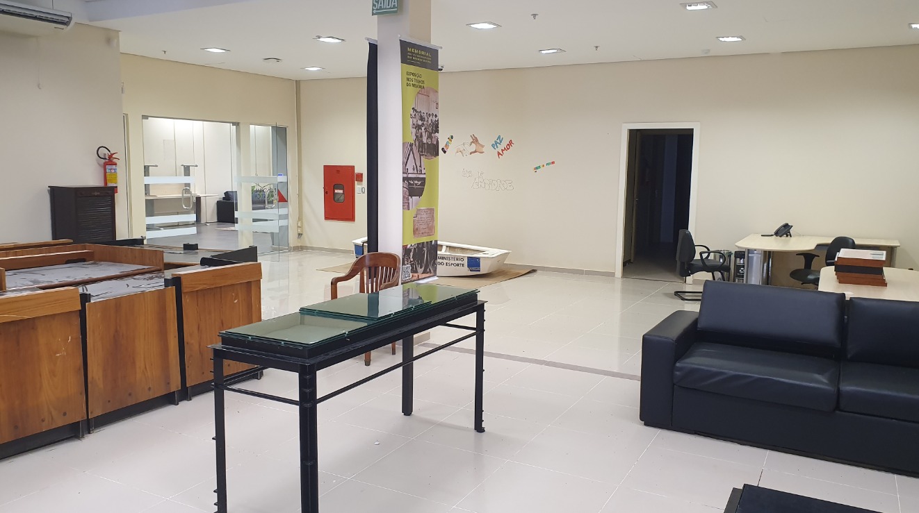 Foto geral da sala, com bancadas de madeira na esquerda, o barco ao fundo, a porta de vidro no fundo à esquerda, uma mesa ao centro, uma porta no centro ao fundo e um sofá na direta.