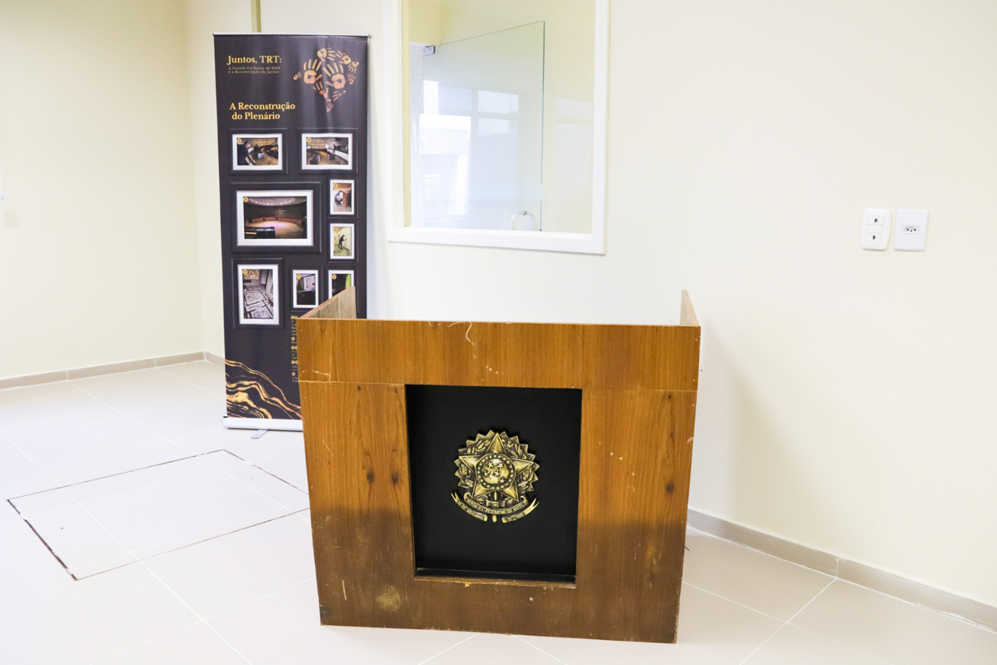 Bancada de madeira com o brasão da República. Ao fundo aparece um cartaz com fotos da enchente e o título "Juntos, TRT"