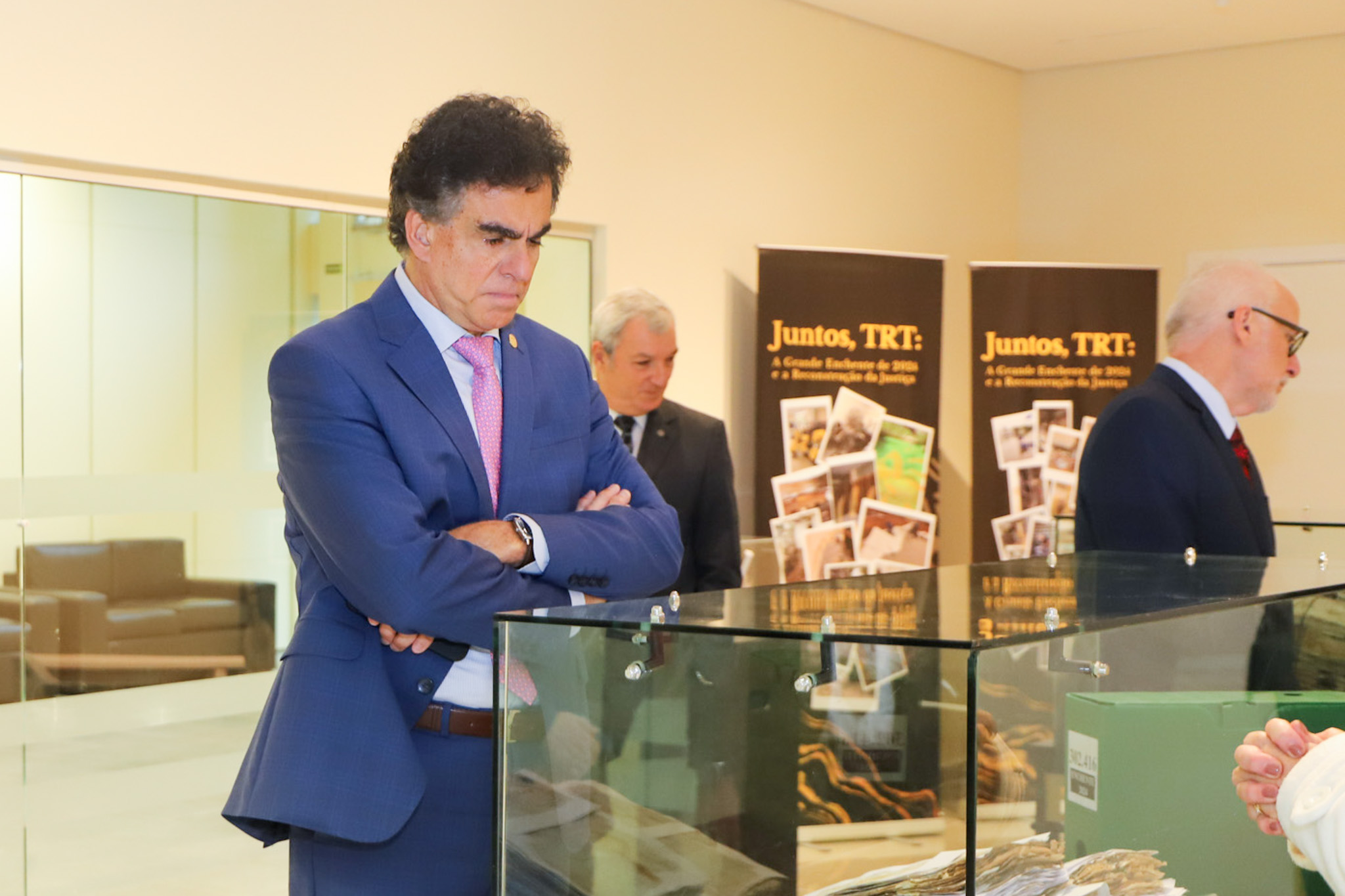 Luiz Philippe está de pé, de braços cruzados, olhando objetos expostos em uma mesa com vitrine. Ele é branco, tem cabelo escuro. Usa terno azul marinho, camisa branca e gravata rosa. 