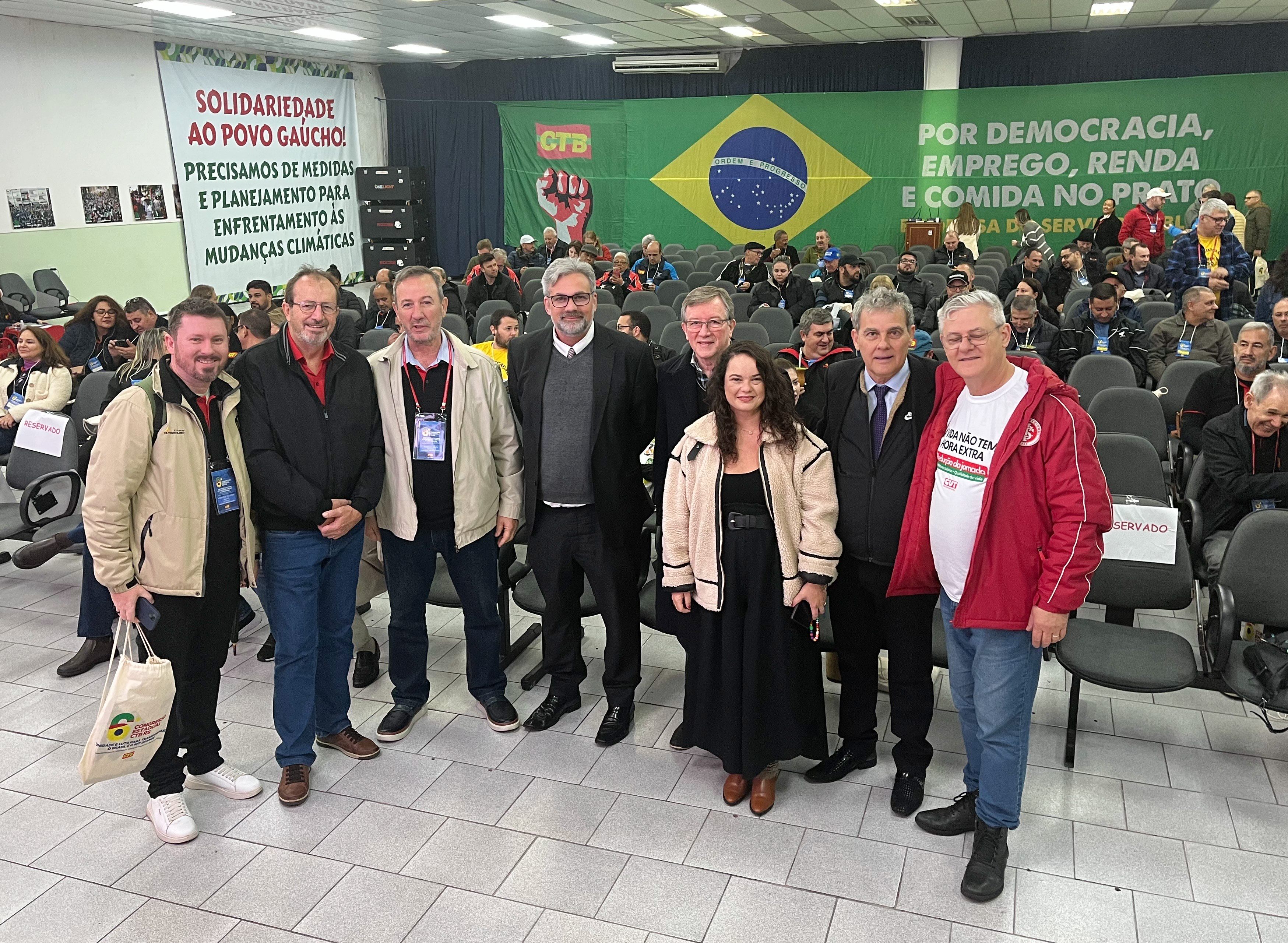 Grupo de sete pessoas, entre elas o desembargador Fraga, posa sorridente em auditório com faixas da CTB ao fundo e público sentado nas cadeiras.
