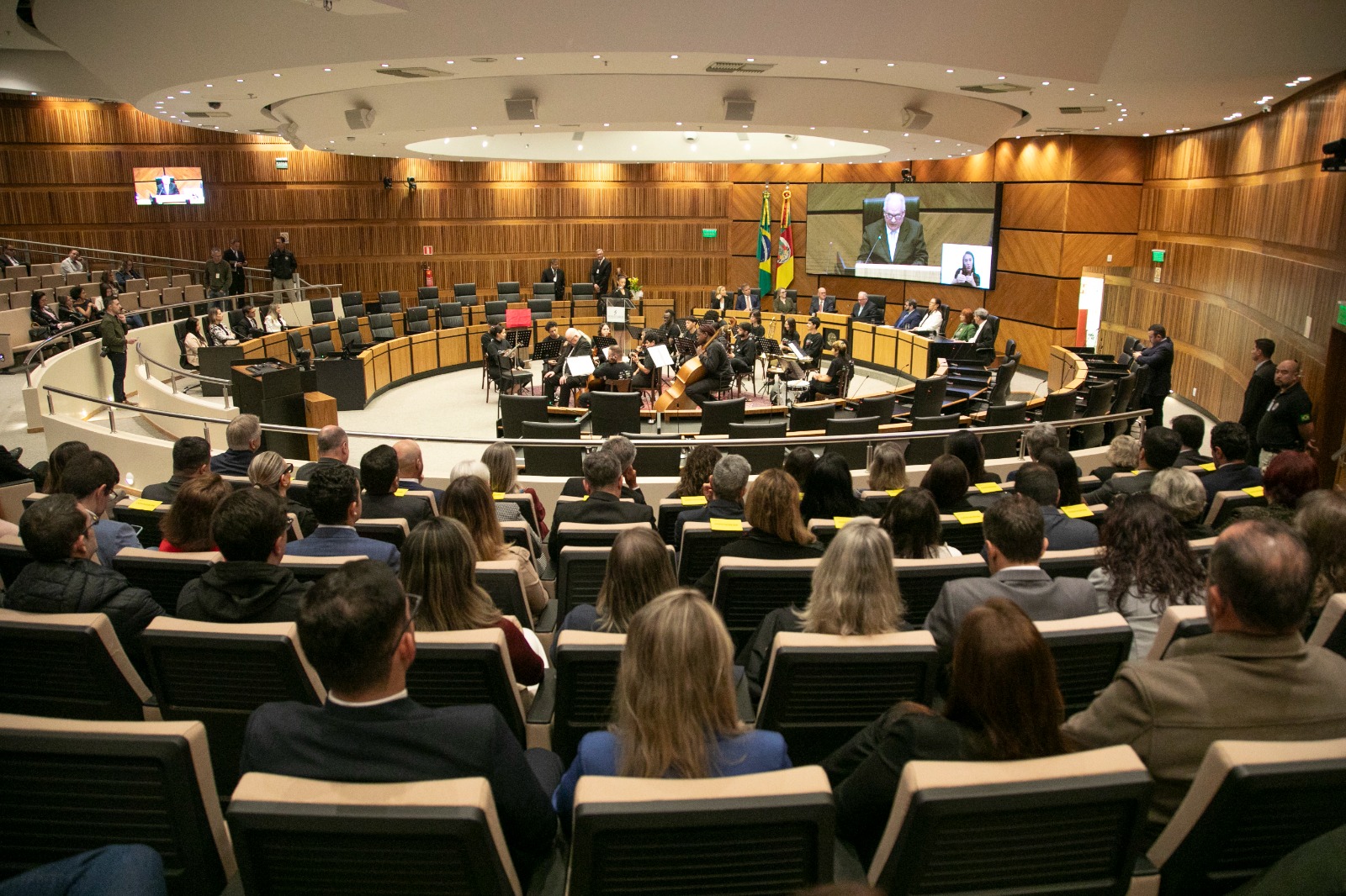 Foto geral do Plenário do TRT-RS, com o público em primeiro plano. Ao fundo, um orquestra se apresenta na nave central. A mesa oficial está no lado direito, e a o telão, também no canto direito, mostra a imagem do ministro Godinho.