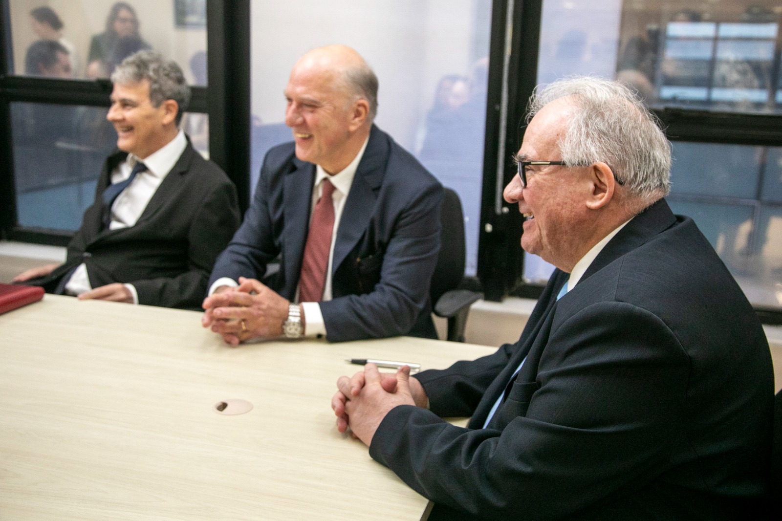 Fraga, Martins Costa e Godinho aparecem sentados na mesa de negociação, sorrindo. Os três são brancos e usam terno e gravata. O ministro Godinho aparece em destaque, no canto direito da foto.