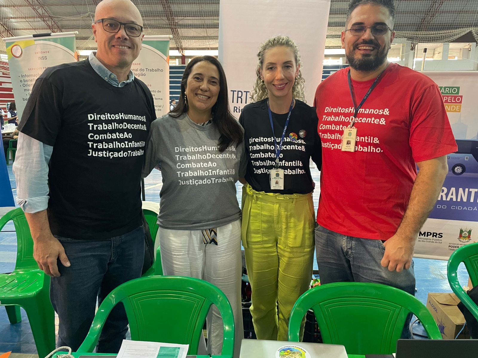 Quatro pessoas (dois homens e duas mulheres) estão sorrindo e posando para a foto. Eles vestem camisetas com frases sobre direitos humanos, trabalho decente e combate ao trabalho infantil. Estão em um ambiente coberto, com cadeiras verdes de plástico ao fundo e banners institucionais do TRT-RS.
