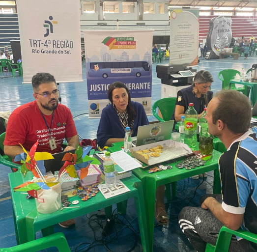 Equipe da Justiça do Trabalho está atendendo um homem em um guichê. Há três pessoas sentadas atrás de uma mesa com papéis, bebidas e materiais informativos. Ao fundo, há faixas e banners do TRT-RS.
