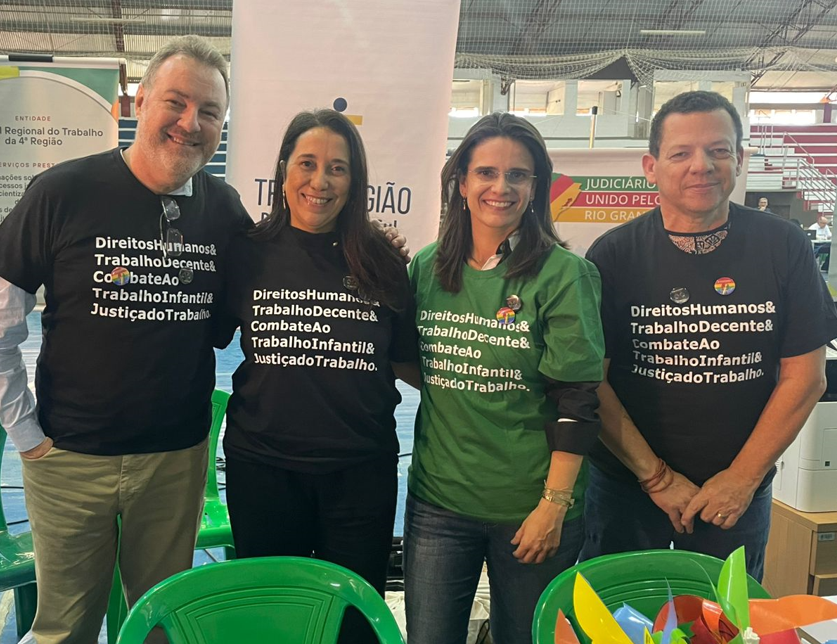 Mais quatro pessoas (dois homens e duas mulheres) estão sorrindo e posando para a foto. Eles vestem camisetas com frases sobre direitos humanos, trabalho decente e combate ao trabalho infantil. Estão em um ambiente coberto, com cadeiras verdes de plástico ao fundo e banners institucionais do TRT-RS.
