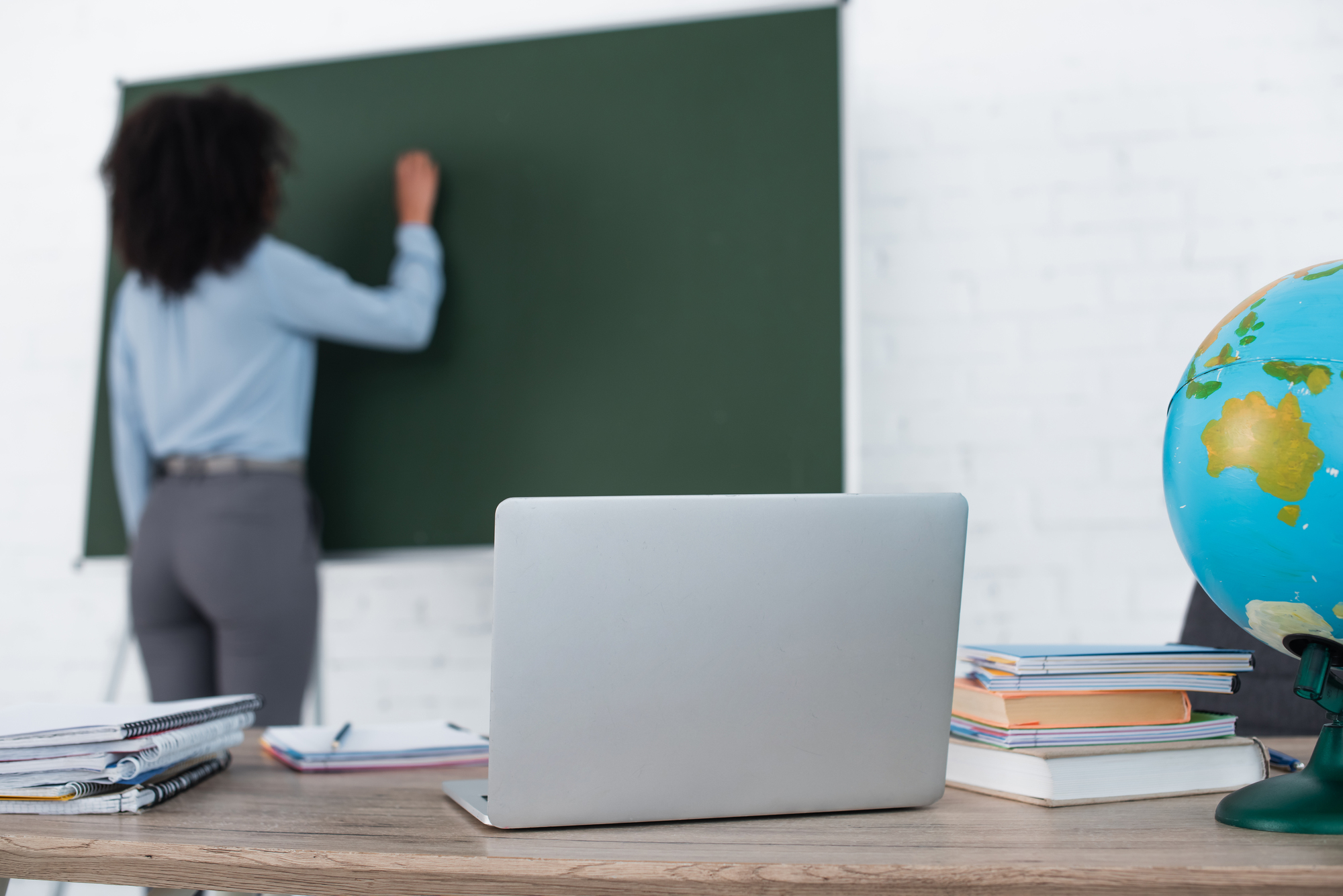 Professora escreve em lousa, na sala de aula. Ela está virada para o quadro e não há imagem do rosto. É uma mulher jovem, magra, negra, com cabelos pretos e crespos. Usa camisa jeans e calça cinza.