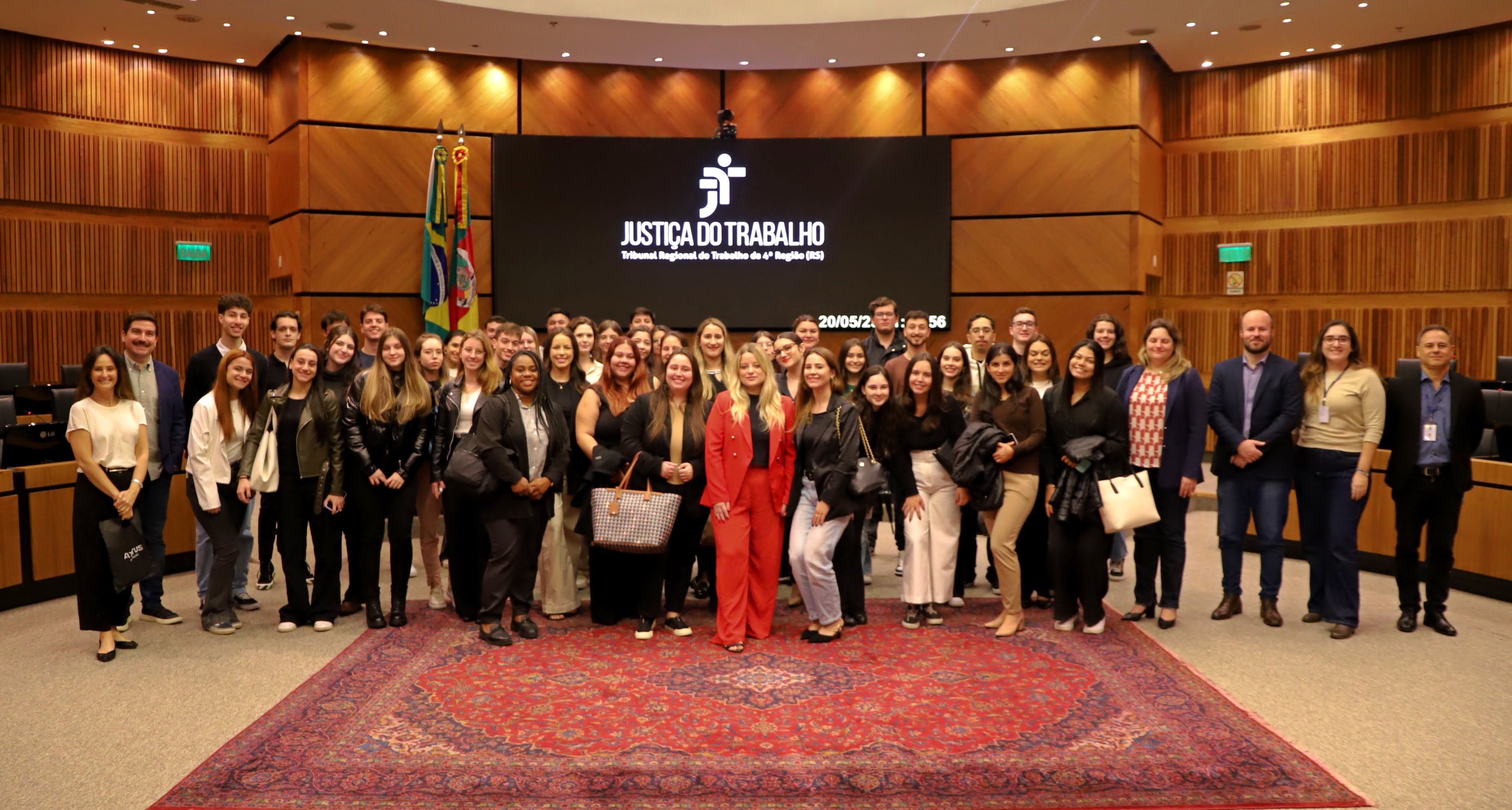 Grupo de aproximadamente 50 pessoas posando para foto em plenário do Tribunal Regional do Trabalho da 4ª Região (RS), com letreiro ao fundo escrito “Justiça do Trabalho”. A maioria são jovens, acompanhados por alguns adultos, sugerindo uma visita institucional ou atividade acadêmica. Ao fundo, bandeiras do Brasil, do Rio Grande do Sul e do tribunal. Todos estão sorrindo e bem-vestidos, com roupas sociais ou casuais elegantes.