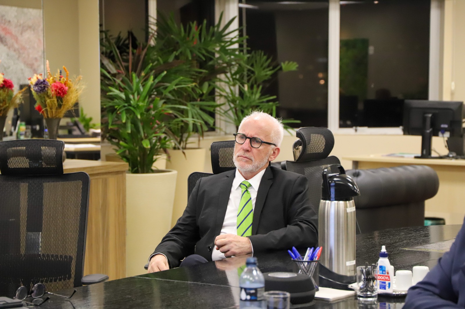 O Juiz Hermann de Araújo Hackradt, de cabelos brancos e barba, usando terno escuro e gravata verde, está sentado à mesa de uma sala de reuniões, com plantas e arranjos florais ao fundo.