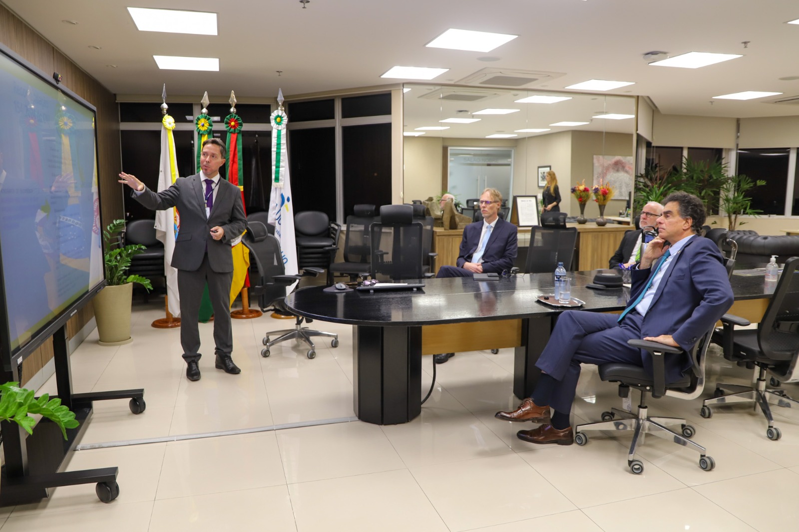 O juiz Rodrigo Trindadefaz uma apresentação diante de uma tela grande, enquanto três participantes sentados à mesa, com bandeiras oficiais ao fundo, acompanham atentamente a exposição.
