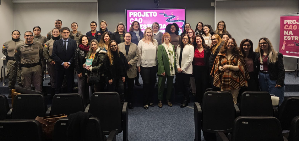 Fotografia com os participantes do evento. A imagem mostra um grupo reunido em um auditório. Ao fundo, é possível ver uma tela exibindo a frase “PROJETO CAO NA ESTRADA” e um banner rosa com o mesmo título.