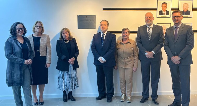 Sete pessoas posam lado a lado em frente a uma parede branca durante a cerimônia de inauguração da galeria de diretoras e diretores da EJERS, no 7º andar da sede histórica do TRE-RS. Ao centro, uma placa comemorativa está fixada na parede, ladeada por quadros com fotos de autoridades. Todos estão vestidos formalmente, demonstrando a solenidade do evento.