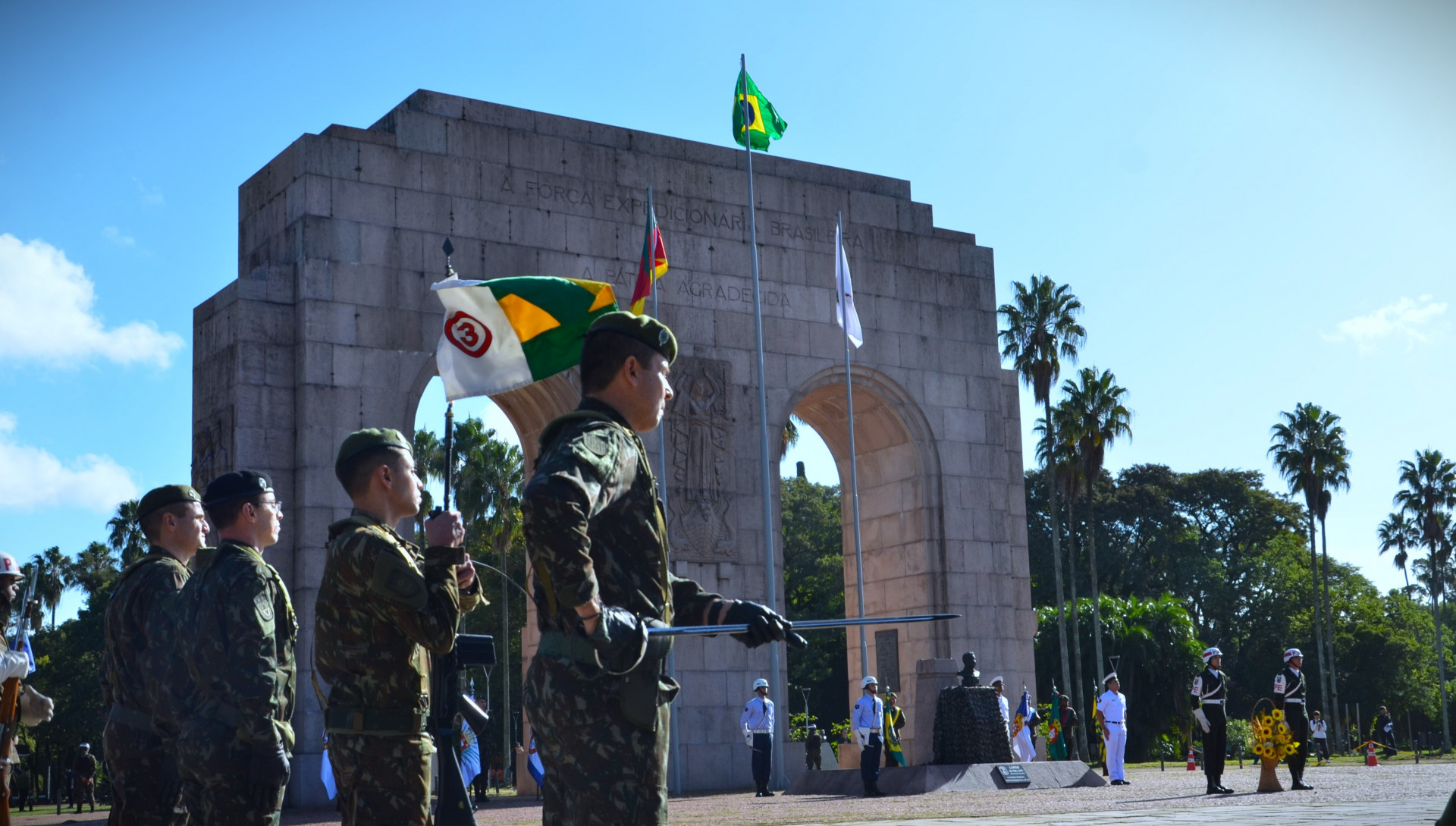 Militares do Exército Brasileiro perfilados prestam continência em frente ao Monumento ao Expedicionário, no Parque Farroupilha, em Porto Alegre, durante cerimônia cívica alusiva aos 80 anos do fim da Segunda Guerra Mundial. Ao fundo, o arco de pedra do monumento exibe a inscrição “A Força Expedicionária Brasileira – A Pátria Agradecida”. Três bandeiras estão hasteadas: a do Brasil, a do Rio Grande do Sul e uma branca.