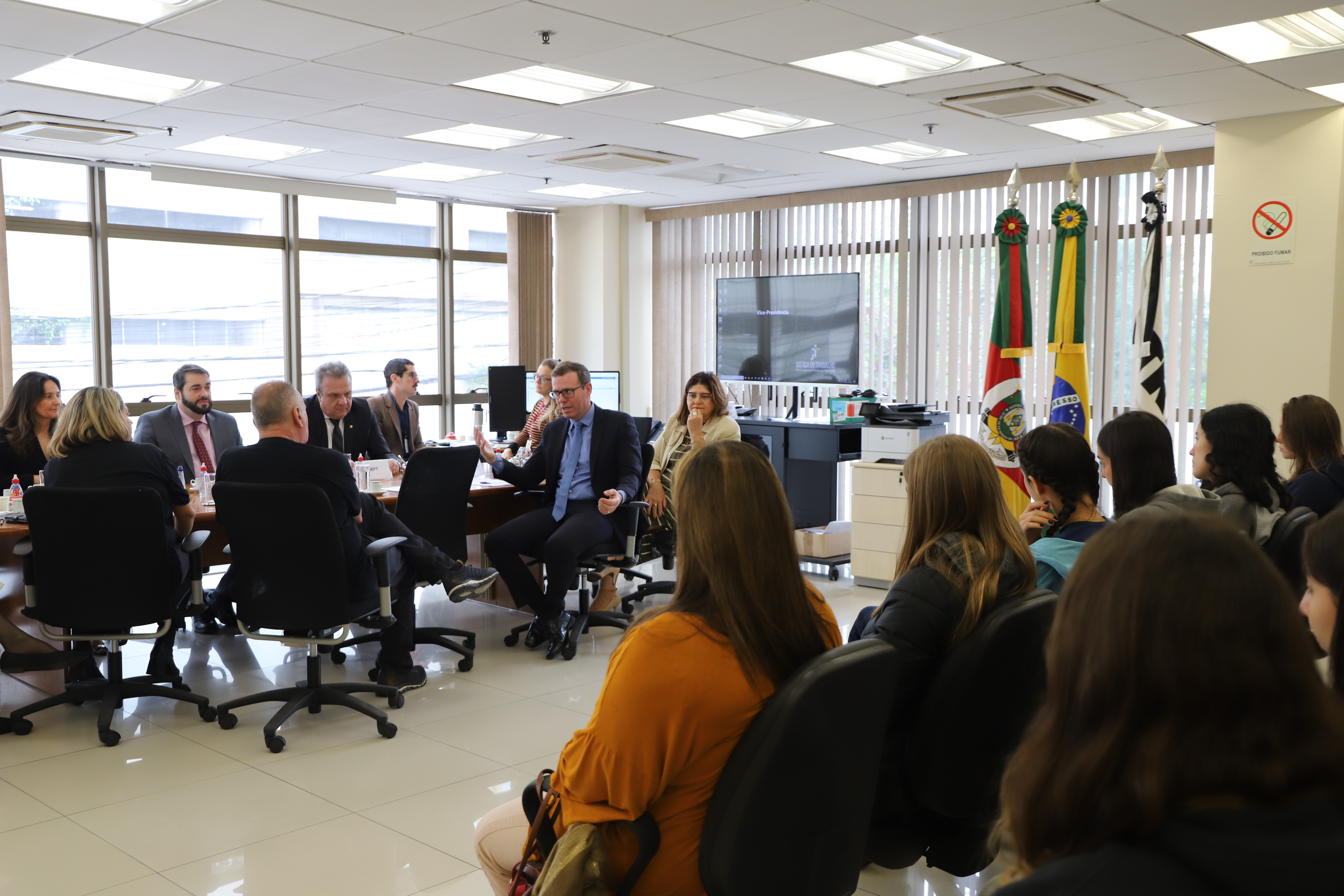 Advogado José, vestindo terno, fala com o grupo de alunos que estão sentados na sala de convivência.