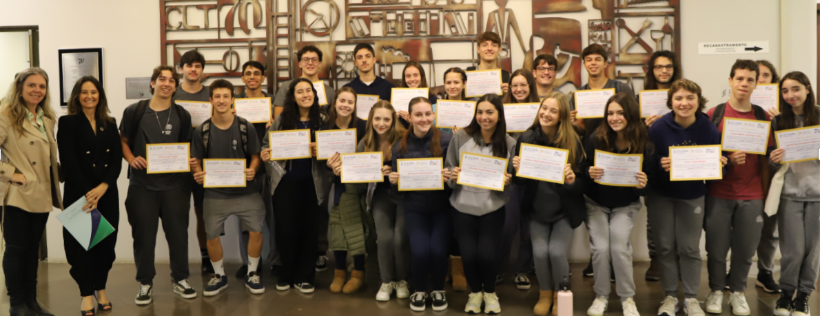Grupo de alunos posando para foto segurando certificados nas mãos. Todos sorriem e estão lado a lado na recepção do Tribunal.