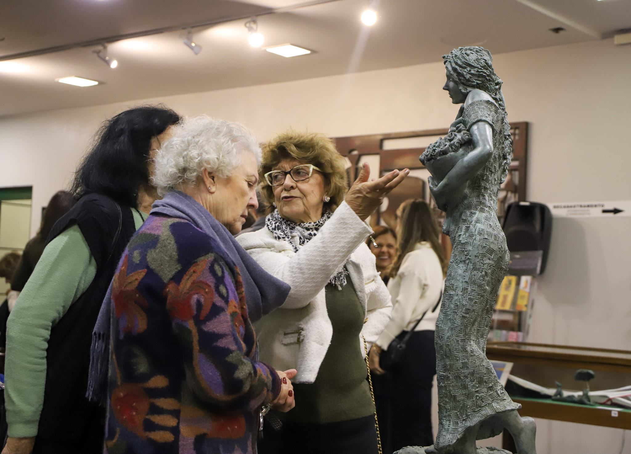 Três mulheres idosas observam atentamente uma escultura de uma figura feminina feita em bronze. Uma delas, de cabelos grisalhos e casaco colorido, olha com interesse; outra, de óculos e casaco branco, gesticula com a mão, como se explicasse algo sobre a obra. A escultura retrata uma mulher em pé, segurando flores junto ao peito. Ao fundo, outras pessoas conversam em um ambiente de galeria ou exposição de arte, bem iluminado.