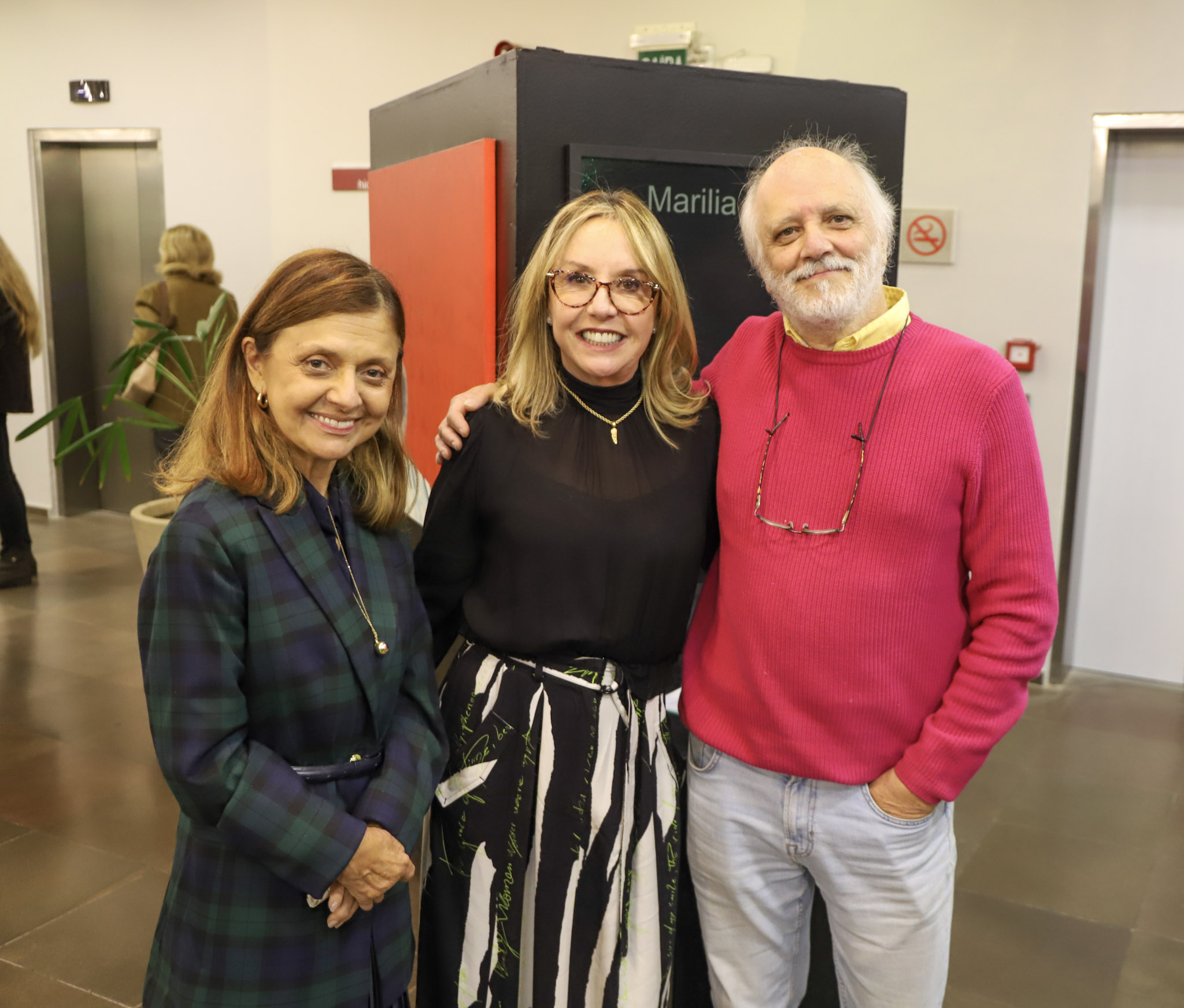 A juíza Anita, a expositora Marilia sorridentes para a foto. Anita usa um casaco xadrez verde-azulado, Marilia veste blusa preta com saia estampada, e o homem, de cabelo branco e barba, está com um suéter rosa e calça jeans. Ao fundo, há uma placa com o nome “Marilia” e portas metálicas.