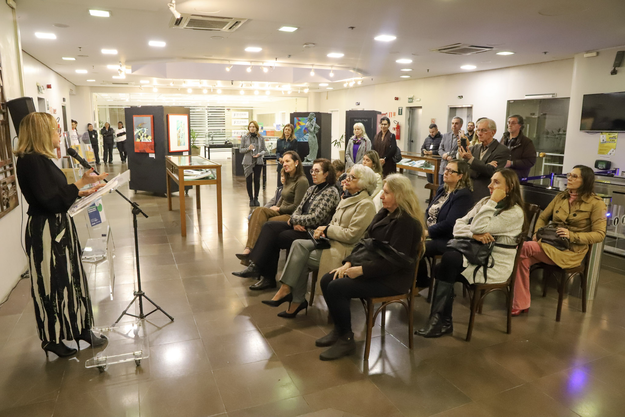 A expositora Marilia fala ao microfone durante a abertura de uma mostra de arte, posicionada ao lado de um púlpito transparente. À sua frente, um grupo de pessoas sentadas, majoritariamente mulheres idosas, ouve atentamente. Ao fundo, outras pessoas em pé acompanham a apresentação e registram o momento com celulares. O ambiente é uma galeria iluminada, com obras expostas nas paredes e vitrines centrais.