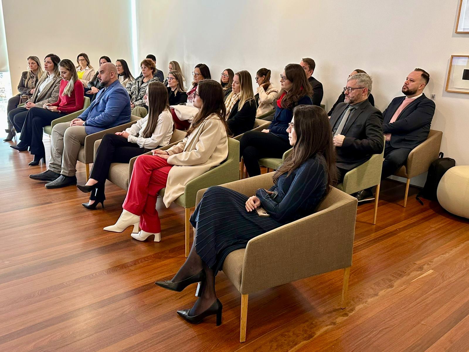 Pessoas assistem a uma apresentação sentadas em cadeiras organizadas em fileiras, durante o evento. Os presentes vestem trajes formais.