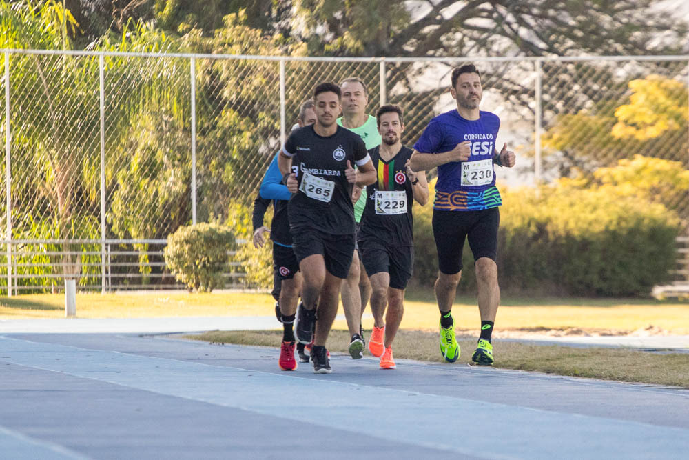 várias pessoas aparecem correndo em um pista atlética azul. Ele usam camisas coloridas com numerações. Ao fundo, aparecem árvores.