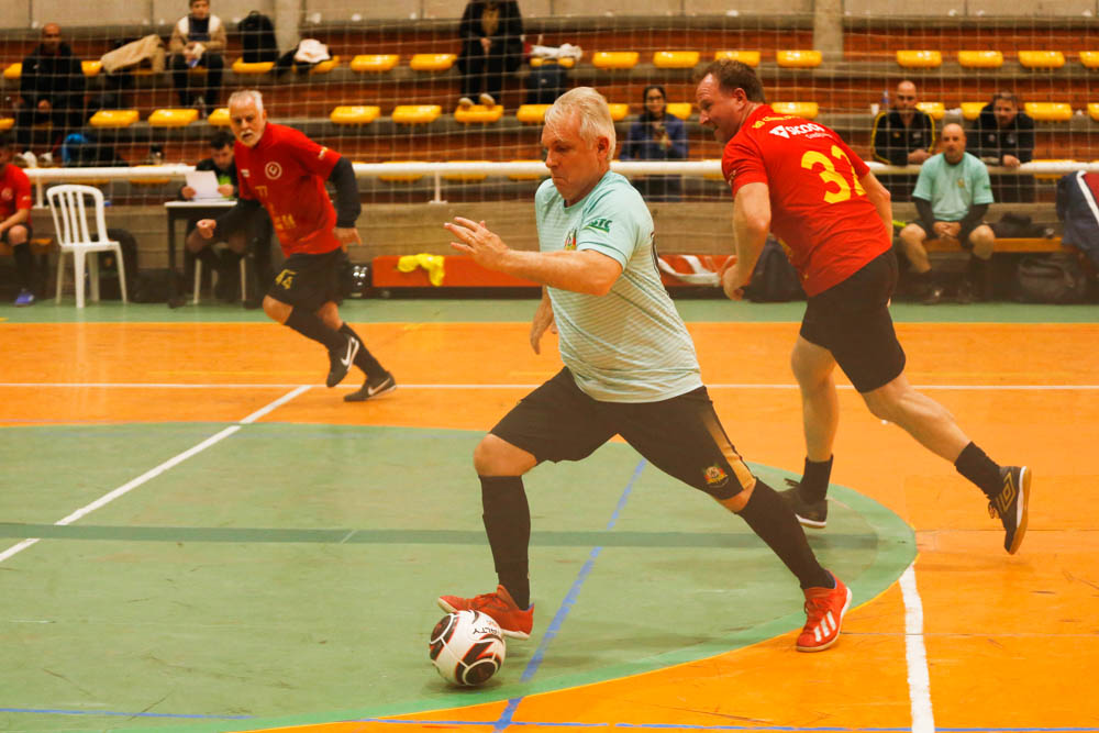 Três homens jogam futsal. Em primeiro plano um deles corre e conduz a bola. Todos correm para o lado esquerdo da imagem. 