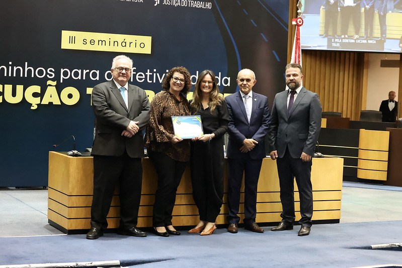 Cinco pessoas posam de pé. Duas mulheres ao centro seguram um certificado Ao fundo aparece o painel do semiário