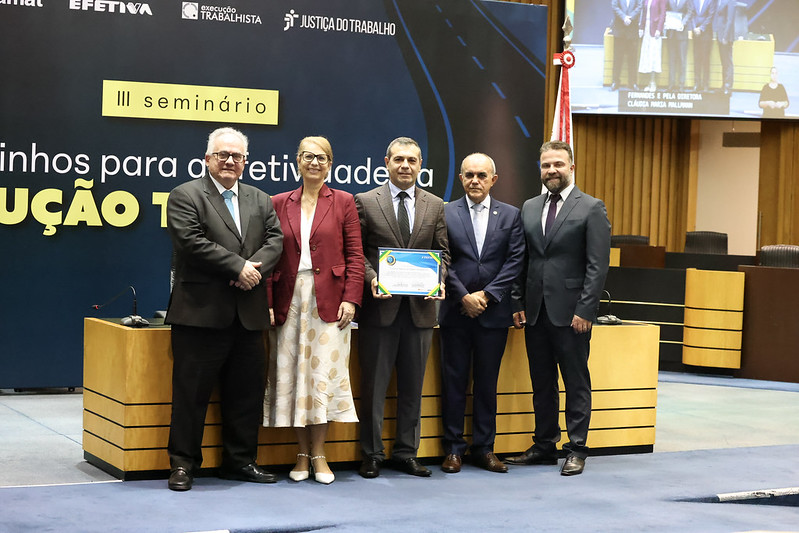 cinco pessoas posam de pé para a foto sorrindo. um homem no centro segura o certificado. ao fundo aparece o painel com o nome do seminário. 