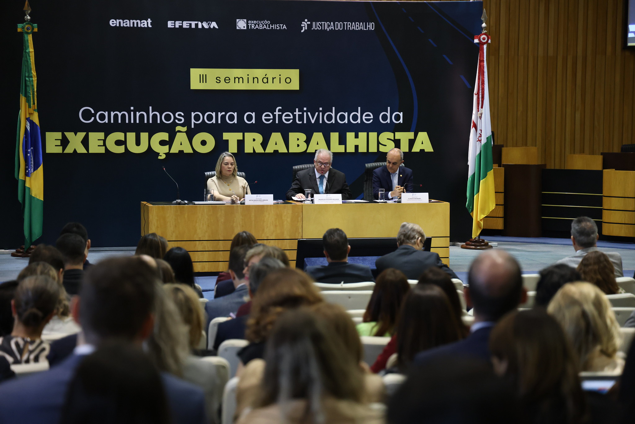 público em primeiro plano. ao fundo, aparecem três pessoas na mesa oficial e um painel atrás com o nome do seminário